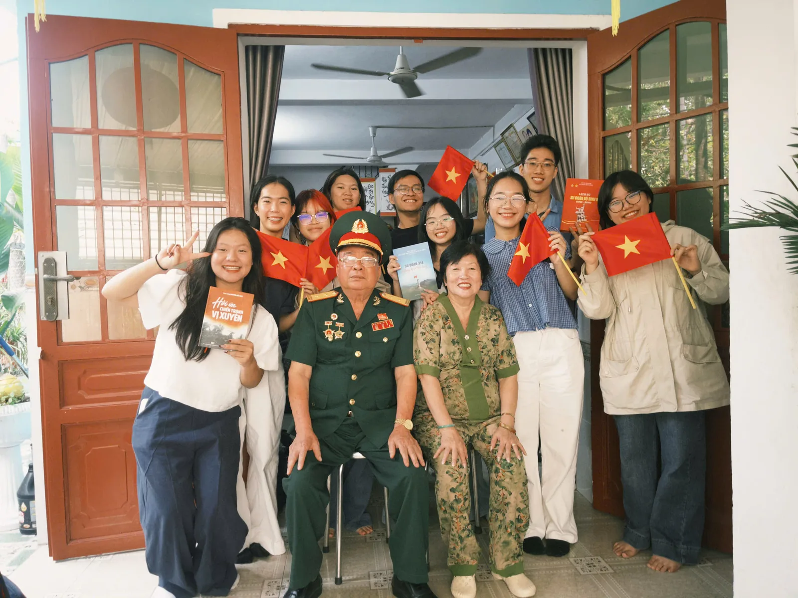 Đoàn lắng cùng một bác cựu chiến binh mặc quân phục và phu nhân, cả nhóm cầm cờ Tổ quốc trước hiên nhà.
