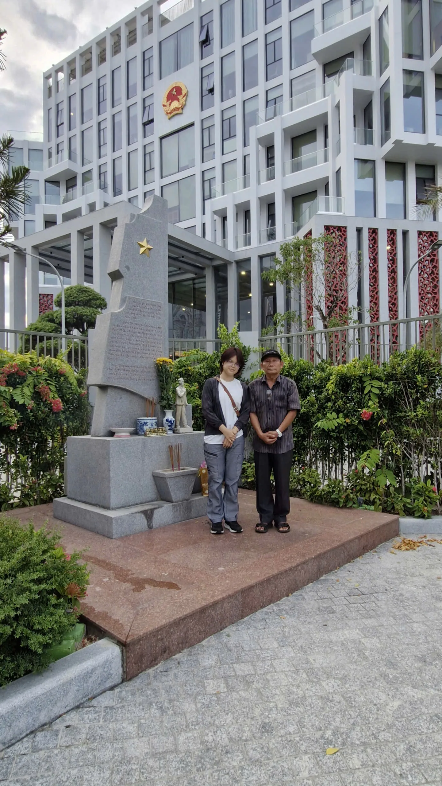 Một bạn trẻ và một bác đứng bên bia tưởng niệm có bát hương đang cháy giữa phố.